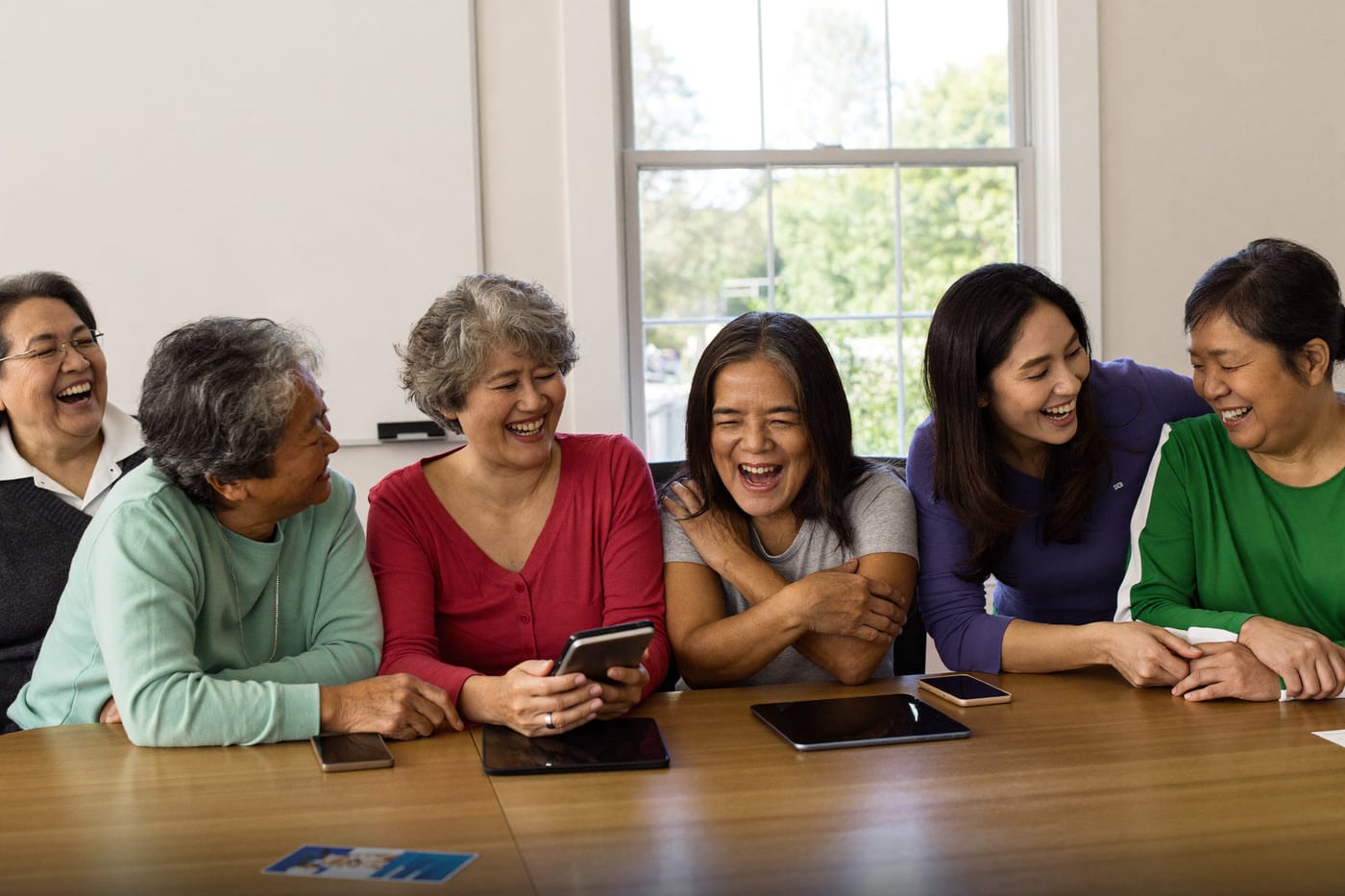 A small group of older adults laugh together around tablets at a community table.