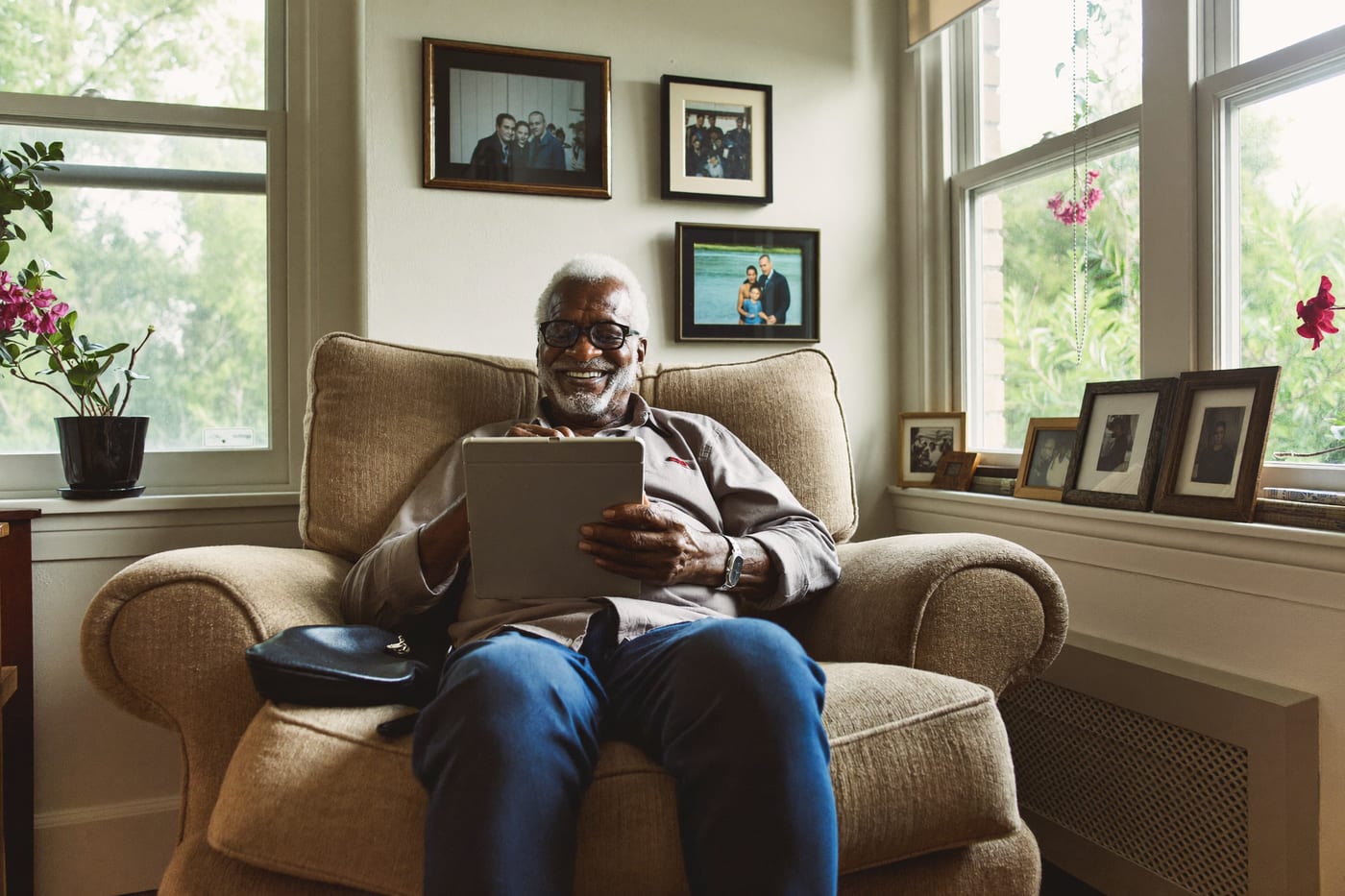 An older man smiles at his tablet in a sunny living room.