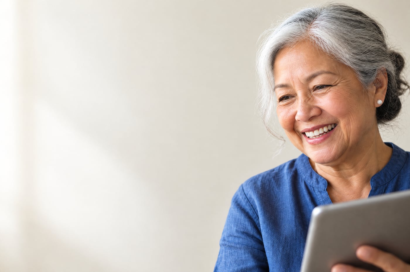 An older Filipina woman smiles warmly at her tablet during a video call with family, in soft daylight.