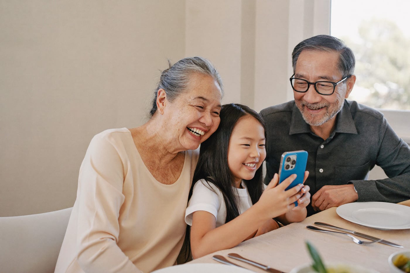 Three generations of a family laugh together around a phone at the dining table.