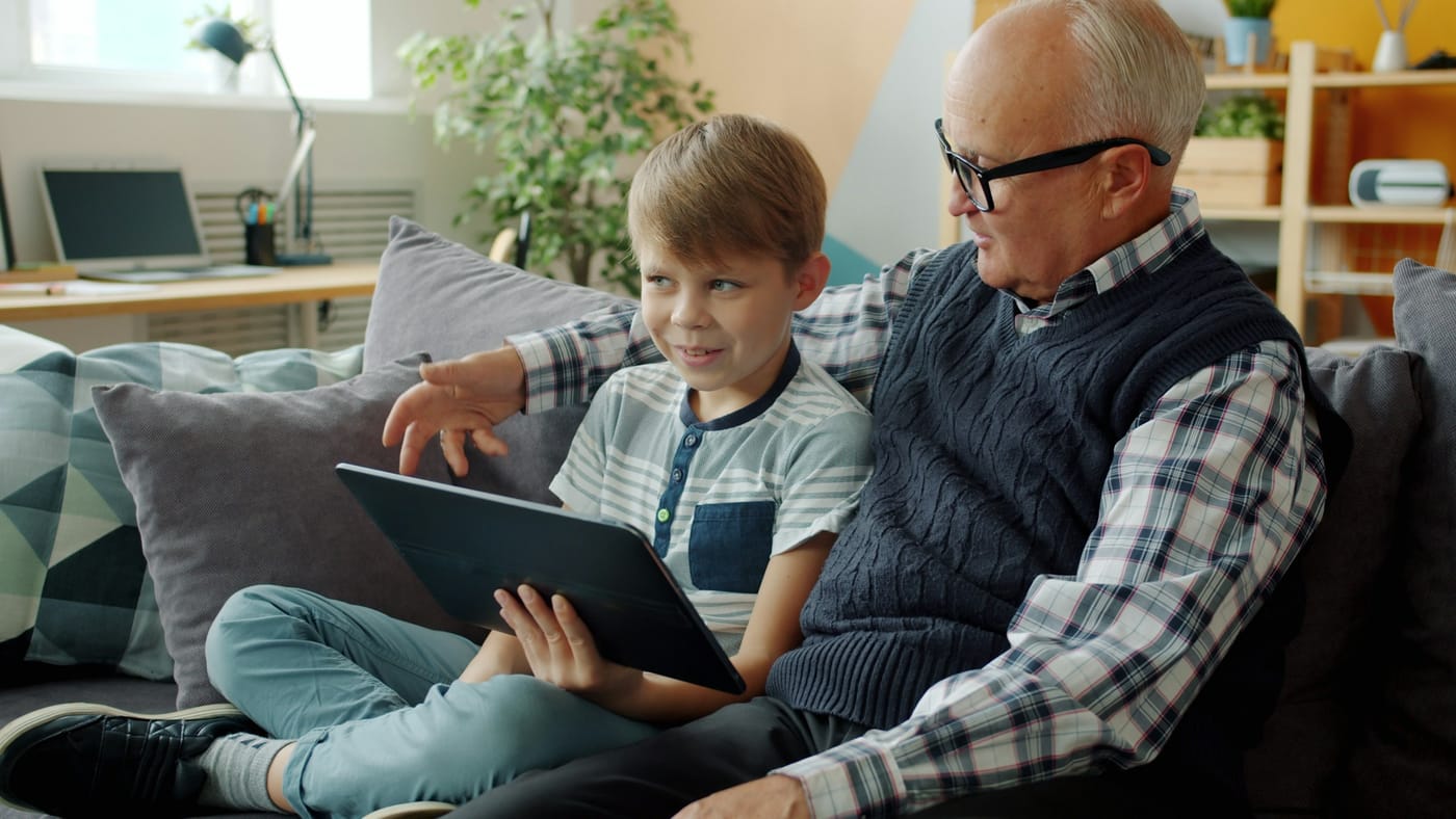 A grandfather and his grandson look at a tablet together on the sofa.